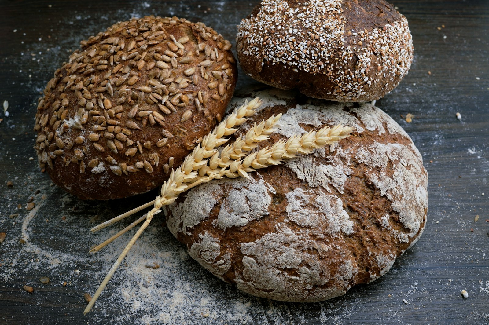 Three round loaves of crusty bread dusted with flour, one topped with seeds, next to a sprig of wheat